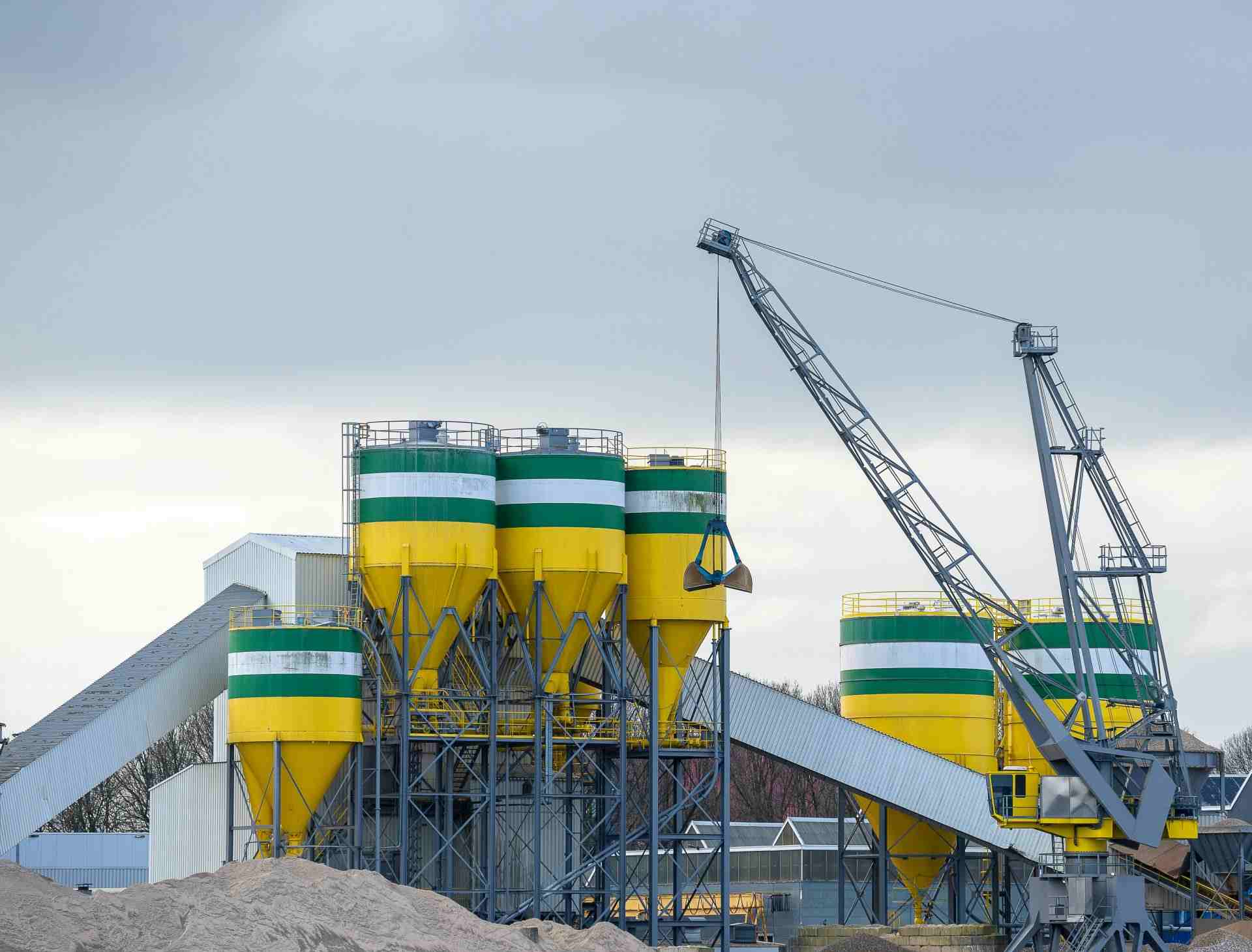 Ein großer Industriekran arbeitet unter bewölktem Himmel mit Silos in einem modernen Zementwerk.
